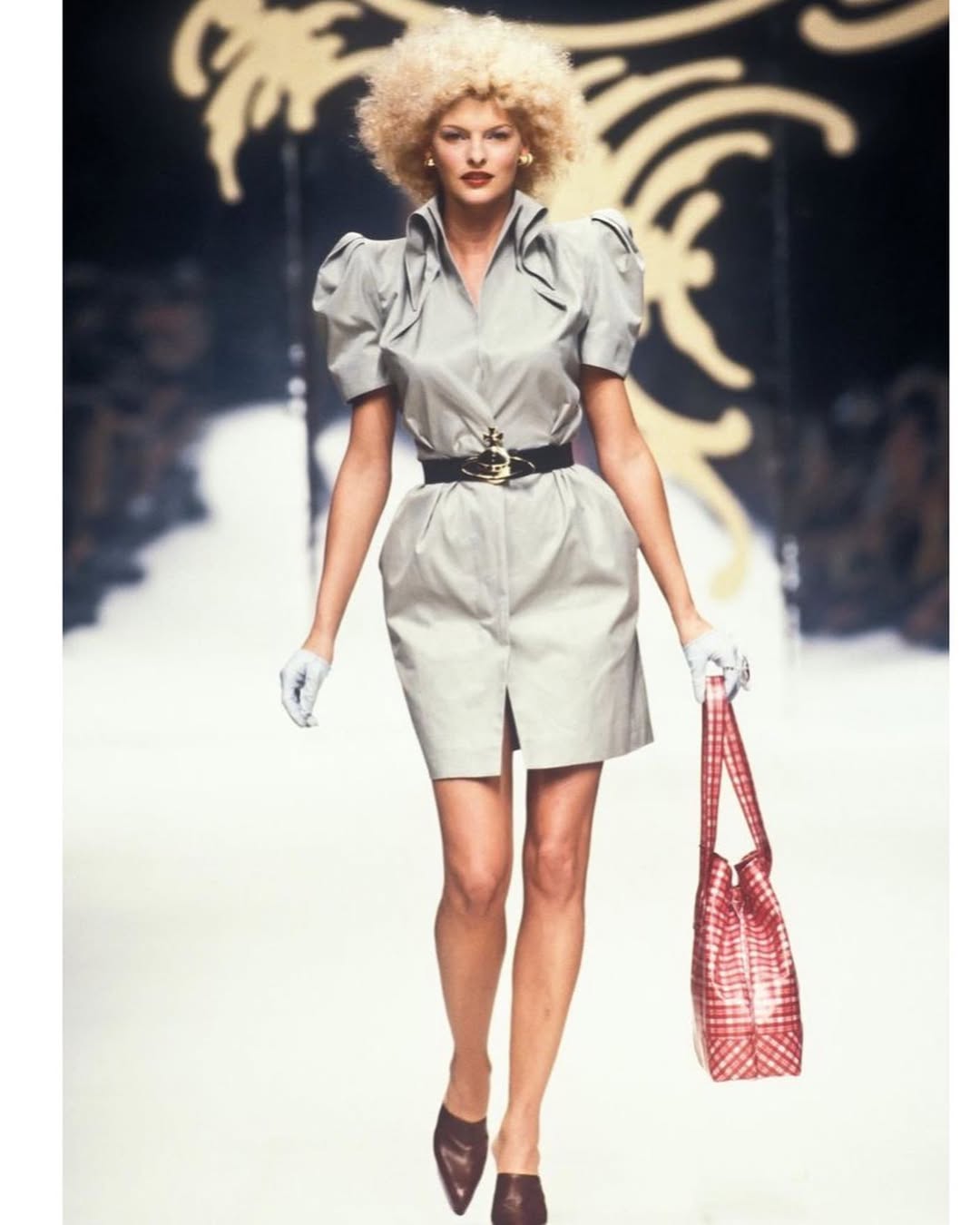 Linda Evangelista is seen wearing a light grey puff-sleeve shirt dress with a belted waist and a red checkered tote bag from Vivienne Westwood’s Spring 1995 “Erotic Zones” collection.