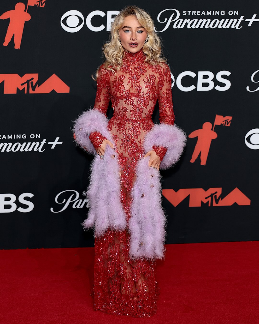 Sabrina Carpenter appears at the 2025 MTV Video Music Awards adorned in an elegant crimson Valentino lace dress complemented by a lavender fur boa.