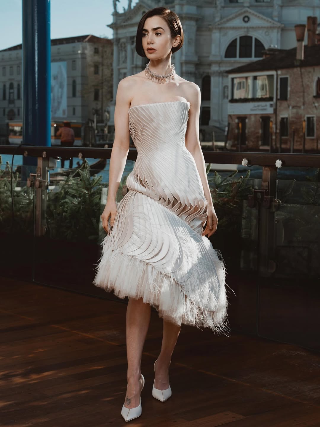 During the “Emily in Paris” NYC Press Tour, Lily Collins wore a strapless ivory Schiaparelli Haute Couture Spring 2025 structured dress with pleated wave accents and a feathered hem.