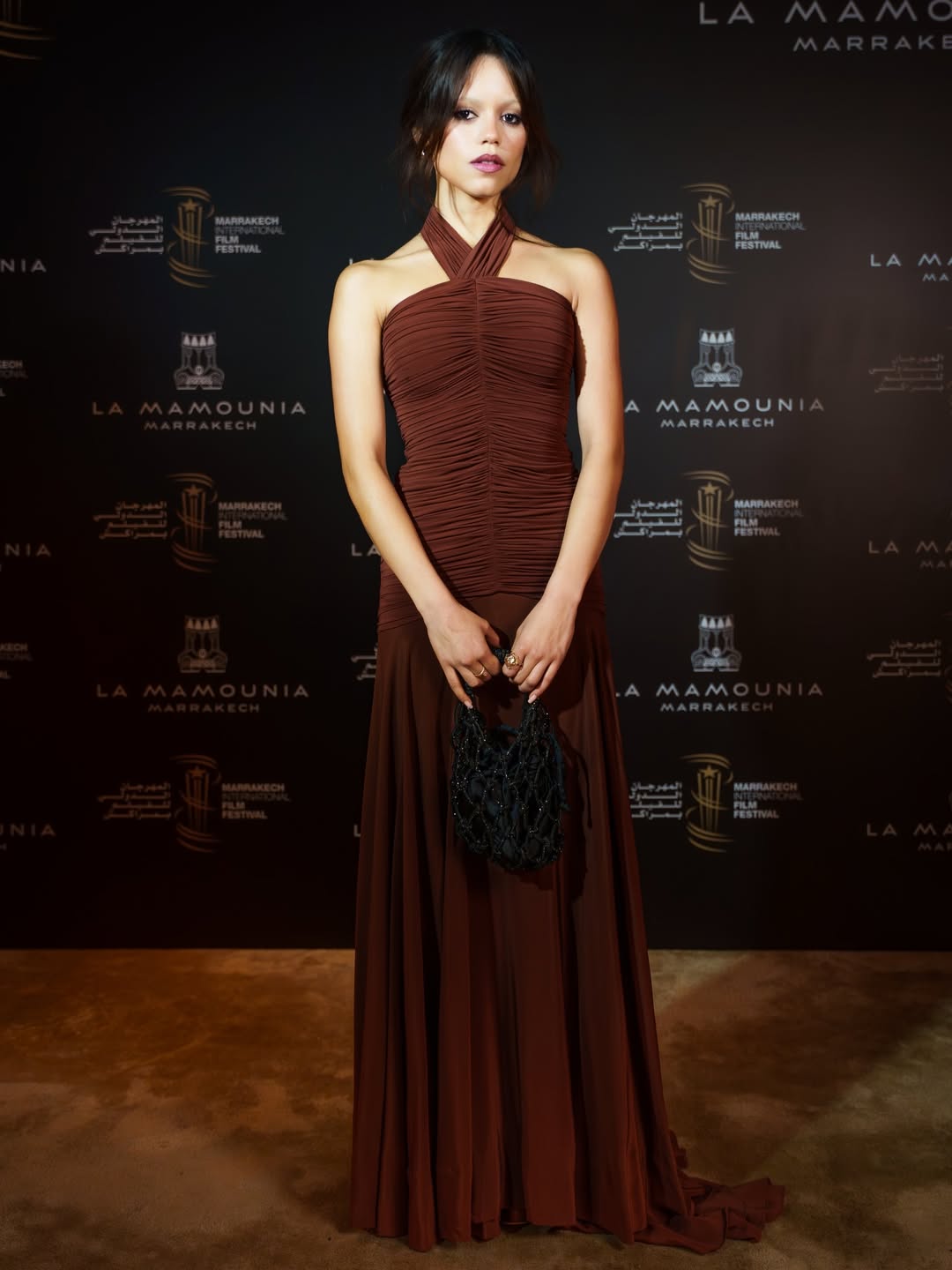 Jenna Ortega wore a sleeveless burgundy Moira silk-blend dress, featuring a unique crisscross halter neck, a gathered bodice, and a flared skirt.