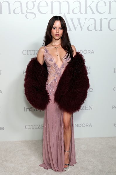 Jenna Ortega in a burgundy Ducie Rochelle shearling coat and a purple sleeveless Amiri dress with a deep V-neck, embroidered bodice, and a thigh-high slit in the floor-length skirt.