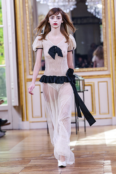 The Enfants Riches Déprimés (ERD) Womenswear Spring/Summer 2026 collection included a cream-white sheer maxi dress with black trim along the short puff sleeves, a black bow detail at the bust, a black ruffled peplum waistline, and an extended black sash tied at the left hip that extended down the side of the dress.
