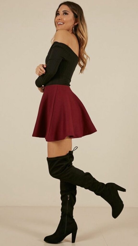 Model in black form fitting off the shoulder long sleeve top with maroon mini skirt and black thigh high boots 