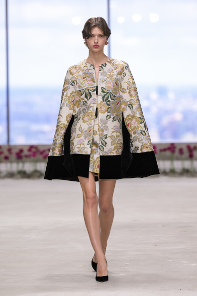 The model is seen striding along the catwalk in an ensemble consisting of a floral and jacquard velvet-trimmed cape paired with a minidress underneath, as showcased in the Carolina Herrera Fall/Winter 2025 collection.