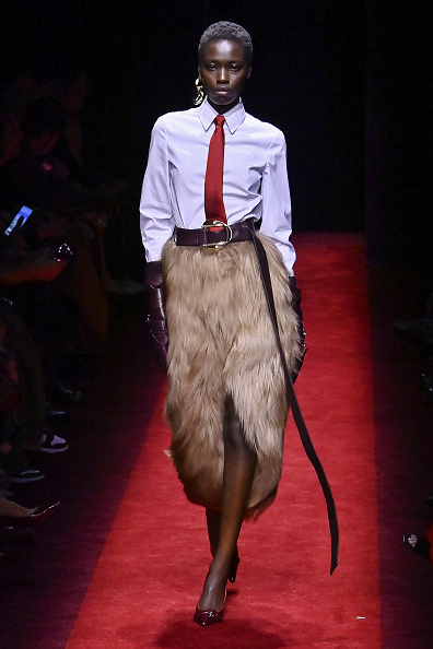 Model Awar Odhiang strides the Ferrari Women’s Fall/Winter 2025/2026 “Officina” ready-to-wear show, donning a long-sleeve collared shirt paired with a tie and a belted fur wrap-around skirt.