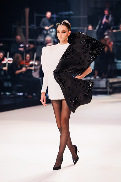 During the Stéphane Rolland Haute Couture Fall/Winter 2025/2026 presentation, a model showcases a white gazar and jersey mini jacket dress featuring long sleeves and adorned with an embroidered black gazar flare.