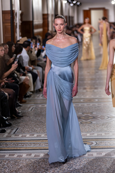 A fashion model walks the runway for Georges Hobeika’s Spring/Summer 2026 collection, dressed in an off-the-shoulder draped satin gown with a corset bodice and sheer chiffon maxi skirt.