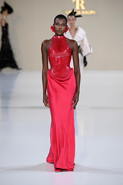 A fashion model walks the Tamara Ralph Haute Couture Spring/Summer 2026 show, “La Lumière Dorée,” wearing a red halterneck dress with a mother-of-pearl corset and a double silk draped skirt.