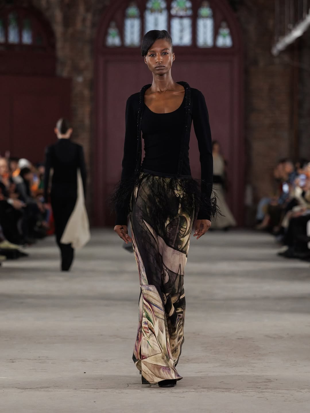 Zahra Traore, a fashion model, is pictured walking down the Diotima Fall/Winter 2026/2027 “Femme Cheval” catwalk wearing a long-sleeved embroidered top and a painted maxi skirt.