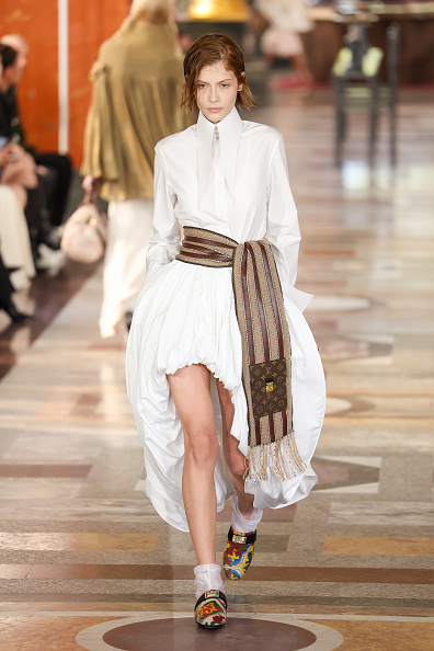 At the Louis Vuitton Women’s Spring/Summer 2026 fashion show, Letícia Hall wore a milky white collared shirt, a striped scarf around her waist, an optic white asymmetrical balloon skirt, and colorful shoes.