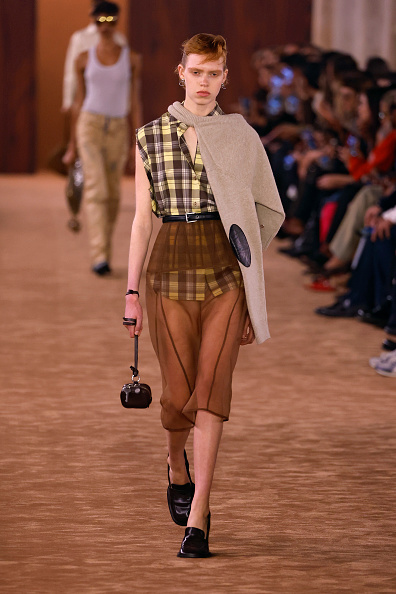 Nanne Groenewegen walked the Acne Studios Women’s Spring/Summer 2026 show wearing a knit sweater draped around her neck and a short-sleeve checkered shirt dress tucked into a high-waisted sheer mesh skirt.