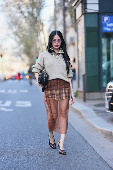 Heart Evangelista wears long black hair, pink tinted sunglasses, a beige knit wool relaxed sweater, a brown and yellow cotton plaid checkered print pattern button down shirt mini dress, a brown sheer mesh pencil midi skirt, a black leather bag, brown leather pumps heels shoes with open toe cap, outside Acne Studios, during Paris Fashion Week