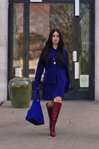 Heart Evangelista wears long black hair, a red cordon with gold pendant necklace, a royal blue suede button-up shirt jacket, a matching royal blue suede knees skirt, a blue suede oversized handbag from Akris, red burgundy leather knee-high block heels boots, outside Akris, during Paris Fashion Week - Womenswear Fall/Winter 2026/2027