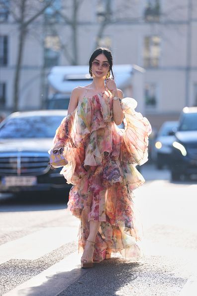 Heart Evangelista wears black hair in a ponytail, pink tinted sunglasses, oversized gold earrings, a white with red / green / pink / blue painted print pattern sheer chiffon floral ruffled dress from Zimmermann, a purple matte leather handbag, beige leather platform high heels shoes, outside Zimmermann, during Paris Fashion Week - Womenswear Fall/Winter 2026/2027