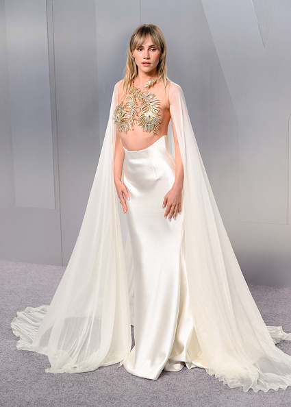 Suki Waterhouse dressed for the 2026 Vanity Fair Oscar Party in a Tamara Ralph Haute Couture sheer silk chiffon cape with a gold crystal peacock feather top underneath, an off-white silk double satin corseted skirt, Stuart Weitzman Nudistcurve Hollywood platform sandals, and Messika Fiery collection-inspired accessories.