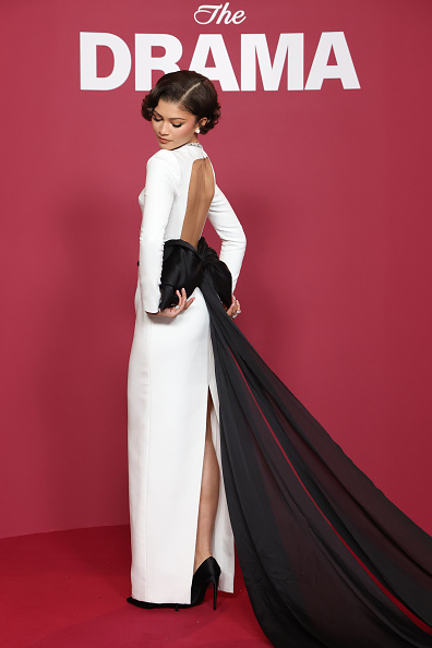 Zendaya walks the red carpet at the Paris premiere of “The Drama” wearing a white long-sleeved Louis Vuitton gown with an open back laced with a long black knotted bow and a high back split that displays her black Louis Vuitton Gala pumps.