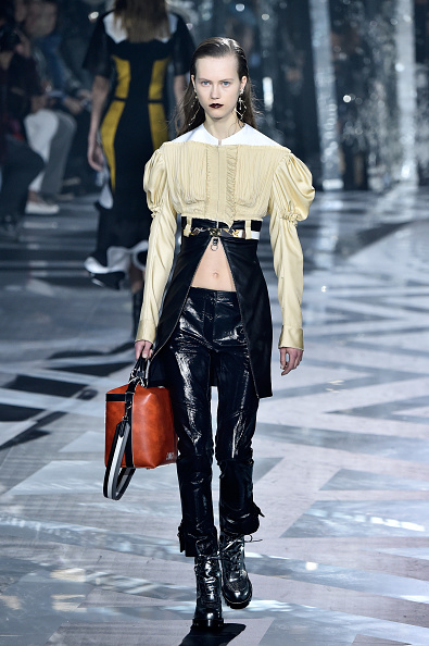 A model strutted the Louis Vuitton Womenswear Fall/Winter 2016/2017 runway, wearing a white pointed-collar shirt, a beige off-the-shoulder puff-sleeve crop top cinched with a strap belt, a black waist-length cloak, and straight-leg leather pants.