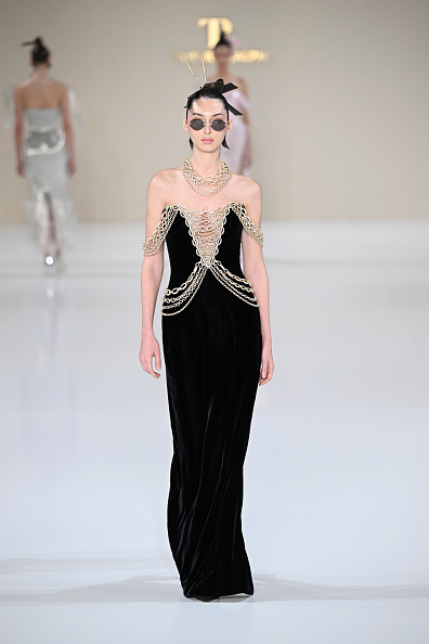 Tamara Ralph Haute Couture showed the “La Lumière Dorée” Spring/Summer 2026 collection during Paris Fashion Week, which included a black off-the-shoulder velvet gown with a gold crystal chain v-cage neckline and gold crystal-draped chains.