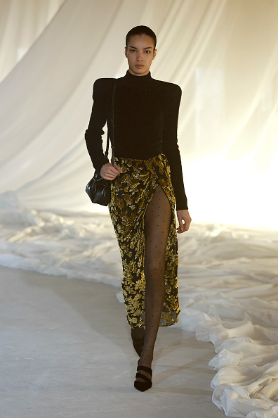 Elise Kouzou walked the runway at the Balmain Womenswear Fall/Winter 2026-2027 ready-to-wear collection fashion show in a turtleneck bodysuit with exaggerated padded shoulders, paired with a high-waisted sheer gold floral jacquard skirt with a thigh-high side slit and sheer polka-dot tights beneath it.