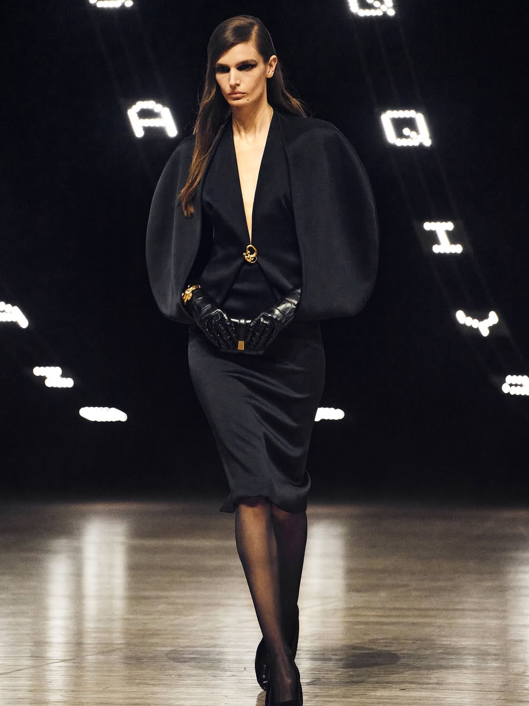 Kate Hudson wore a Khaite silk crepe back satin midi dress from the Fall/Winter 2026 collection to the 2026 Time100 Gala, which featured a plunging neckline embellished with a gold monkey-shaped brooch, sculptural cape sleeves with slits, patent leather cuff gloves accented with an 18 kt gold-plated brass monkey bracelet, and a midi skirt paired with sheer pantyhose.