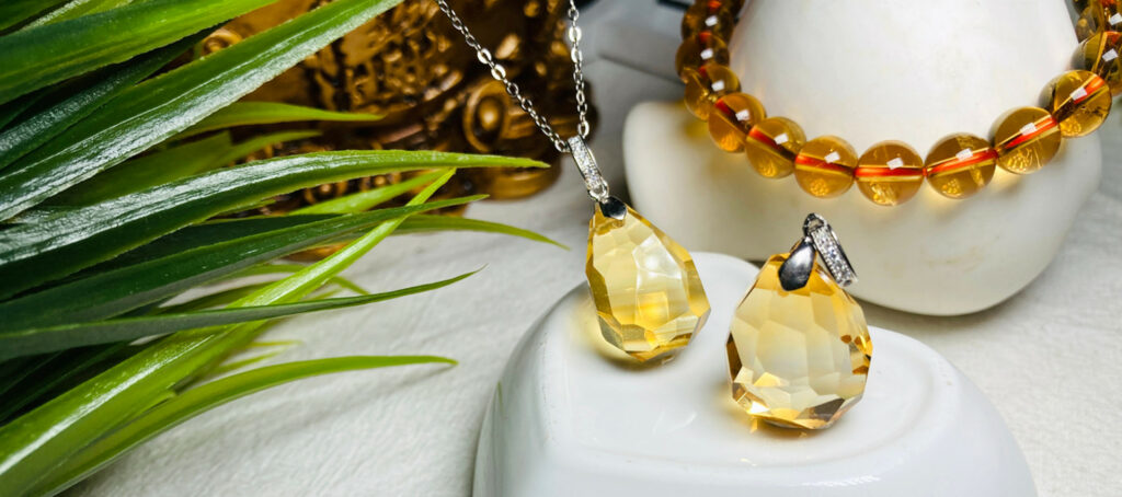 November Birthstone - Faceted yellow teardrop pendant and matching earrings with an amber bead necklace on a white display amid green leaves.