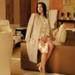 Woman in a textured beige coat over a cream outfit sits on a brown ottoman in a luxe hotel lobby, smiling.