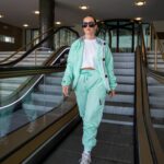 Woman in a mint-green tracksuit descends an indoor escalator, wearing a white crop top and large sunglasses.