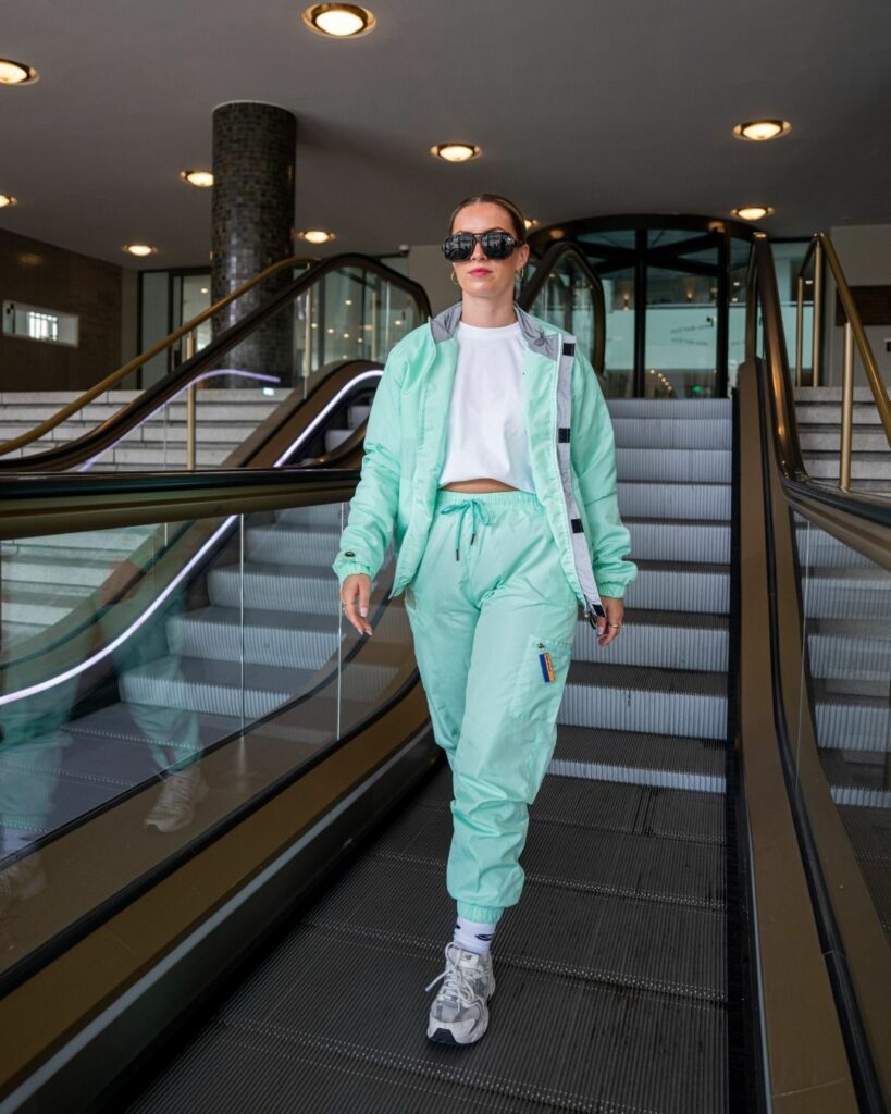 Woman in a mint-green tracksuit descends an indoor escalator, wearing a white crop top and large sunglasses.