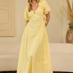 Woman in a pale yellow maxi dress with puff sleeves and a V-neck, standing in a neutral living room setting with a beige sofa and art in the background.