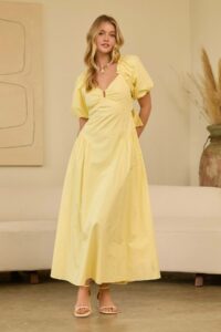 Woman in a pale yellow maxi dress with puff sleeves and a V-neck, standing in a neutral living room setting with a beige sofa and art in the background.