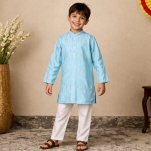 Smiling boy in a light blue kurta and white pants standing indoors with a beige wall background and a plant to the left.
