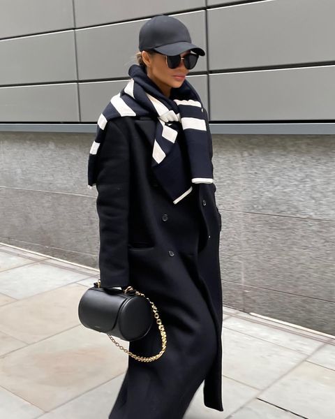 Woman walking outside in a long black coat, striped scarf, black cap and sunglasses, carrying a round black Barrel Bags with a gold chain.