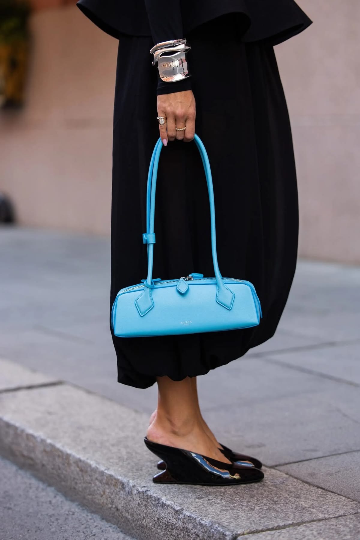Woman in a black dress holding a sky-blue East-West bag by its handles on steps.