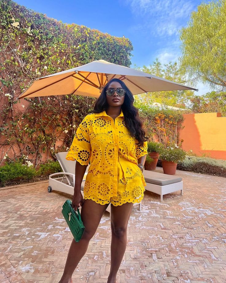 Woman in a bright yellow lace romper standing outdoors, holding a green clutch, wearing sunglasses under a large patio umbrella.
