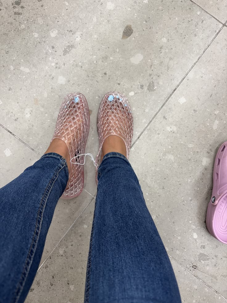 Pair of pink jelly flats worn with blue jeans, standing on a light gray tiled floor; a pink Crocs clog is visible to the right.