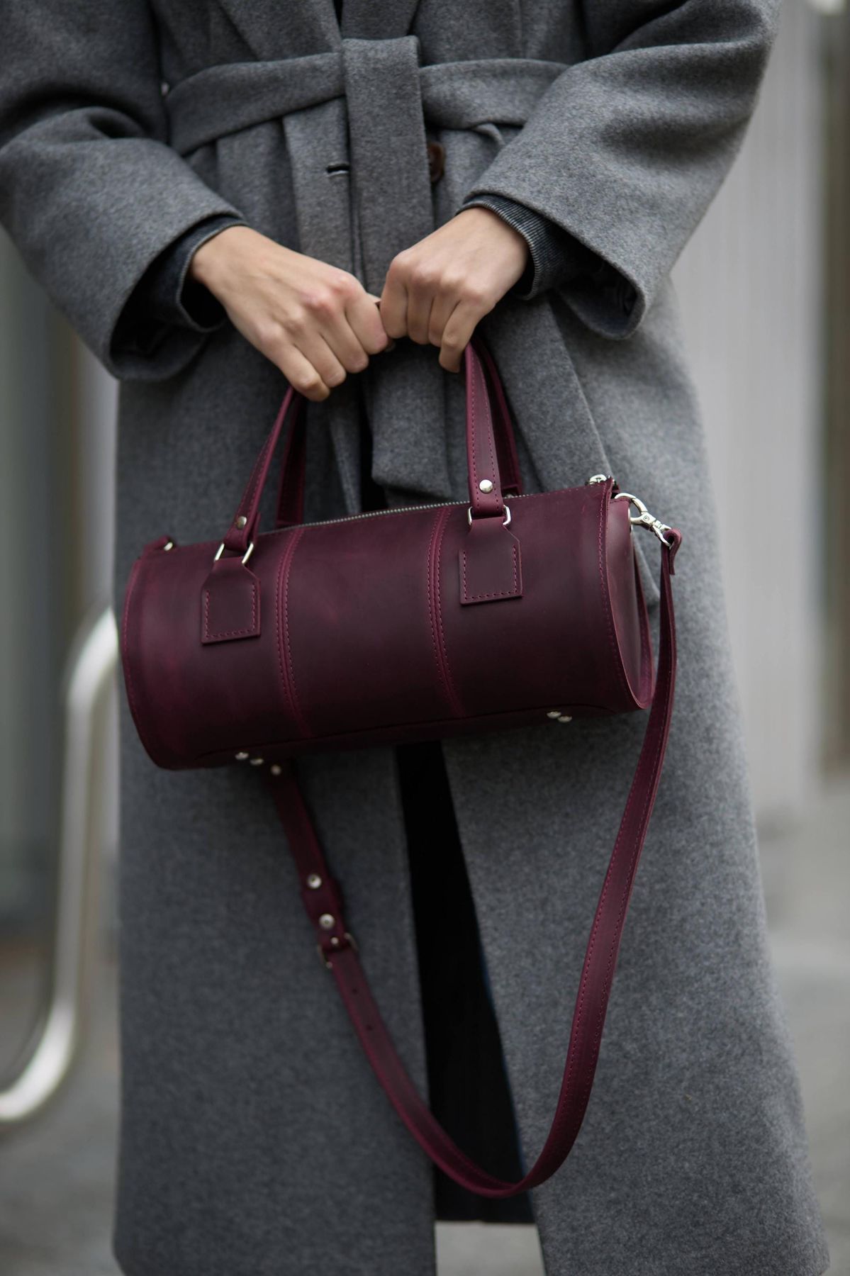 Person in a gray coat holds a burgundy leather Barrel Bags by its handles on a city street.