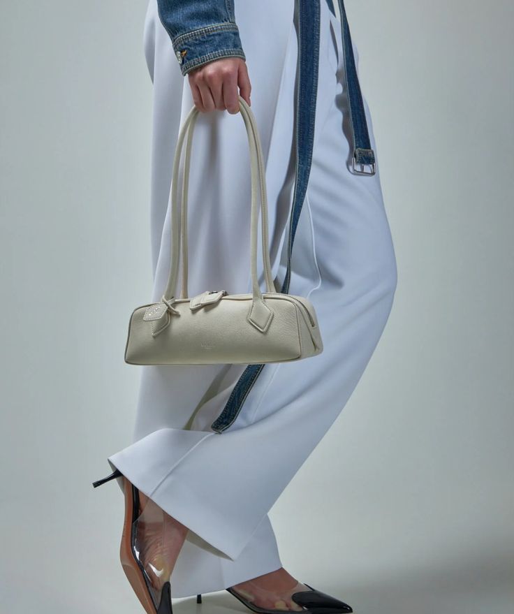 Model holding a small beige East-West bag with double top handles, wearing a white dress and denim jacket.