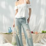 She stands outdoors wearing a white off-the-shoulder top and light-wash ripped jeans, with a table of fruit behind her.