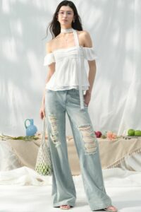 She stands outdoors wearing a white off-the-shoulder top and light-wash ripped jeans, with a table of fruit behind her.