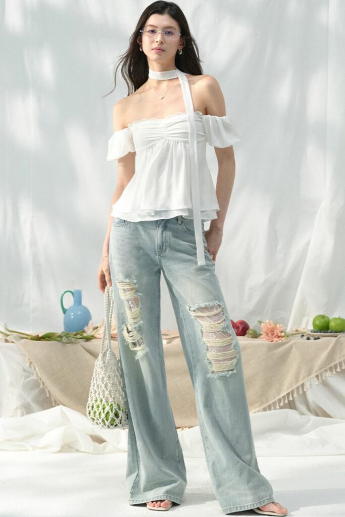 She stands outdoors wearing a white off-the-shoulder top and light-wash ripped jeans, with a table of fruit behind her.