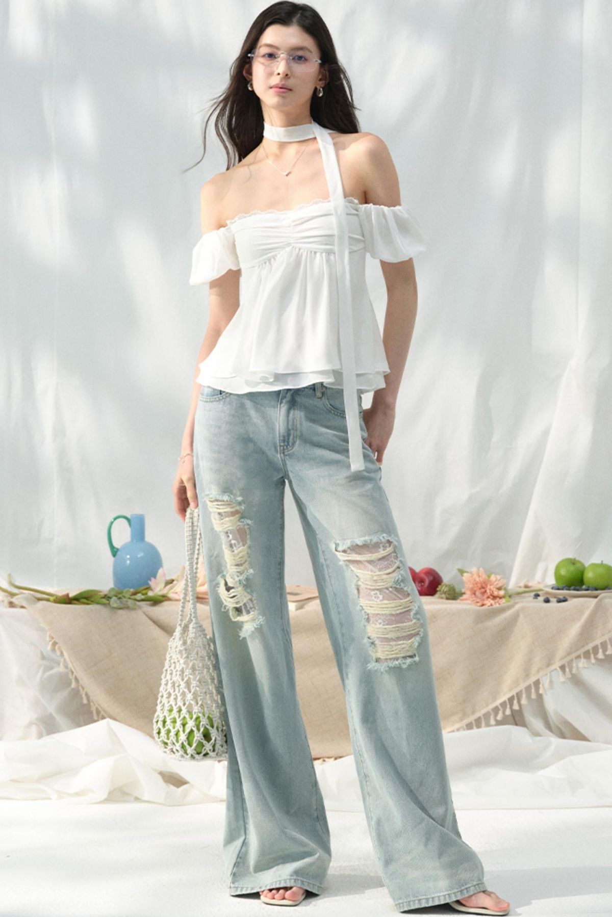 She stands outdoors wearing a white off-the-shoulder top and light-wash ripped jeans, with a table of fruit behind her.