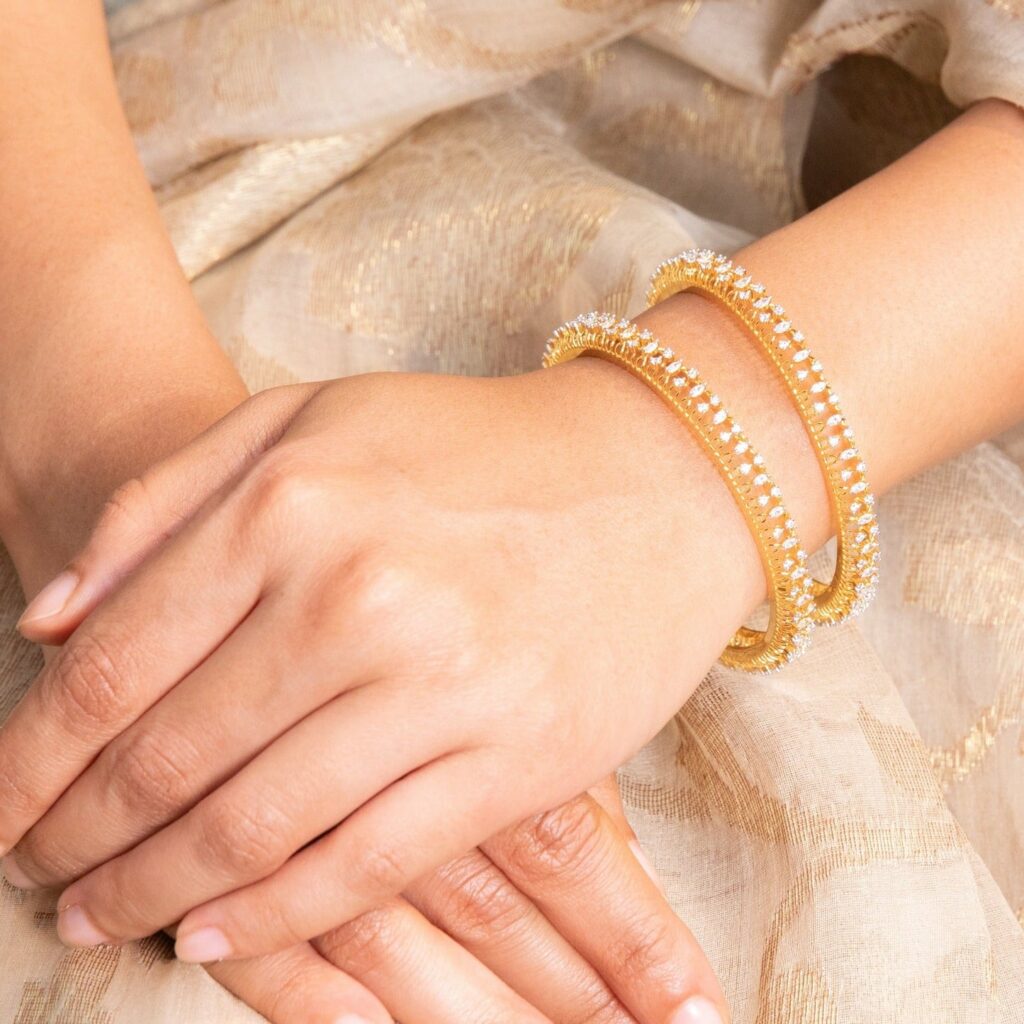 Close-up of two diamond-studded gold bangles on a wrist resting on beige fabric.