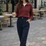 Woman walking on a city sidewalk holding iced coffee, wearing a red pullover, navy trousers, white sneakers, and sunglasses.