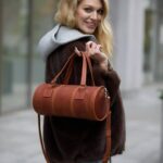 Smiling woman in a dark fur coat with a gray hoodie, carrying a brown leather cylindrical handbag over her shoulder on a city sidewalk.