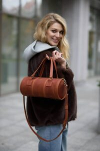 Smiling woman in a dark fur coat with a gray hoodie, carrying a brown leather cylindrical handbag over her shoulder on a city sidewalk.