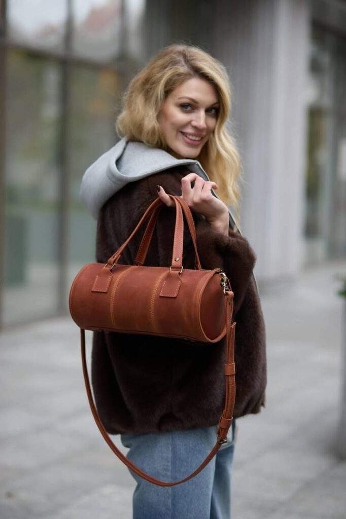 Smiling woman in a dark fur coat with a gray hoodie, carrying a brown leather cylindrical handbag over her shoulder on a city sidewalk.