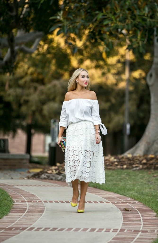 Woman in an off-the-shoulder white blouse and knee-length lace skirt walks along a brick-pathed park path, wearing yellow heels and carrying a colorful clutch.