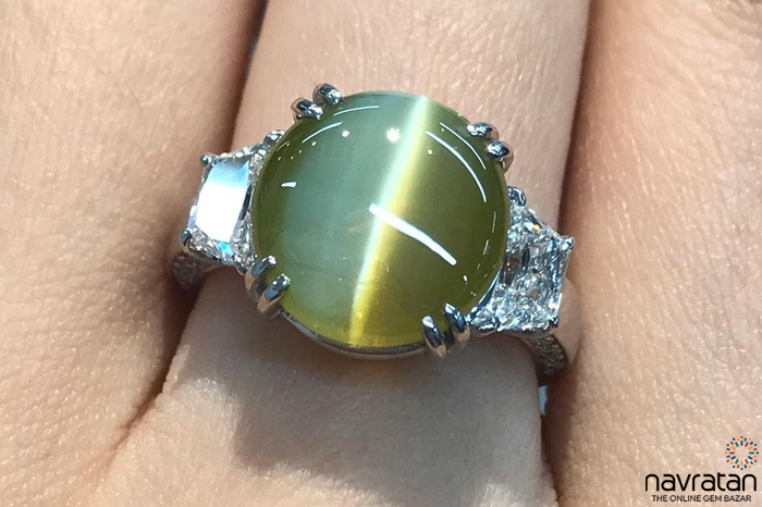 Close-up of a cat's eye ring with an oval green-yellow cabochon gemstone and diamond-accent band on a finger, showcasing the prong setting.