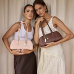 Two women pose side by side, each holding a structured handbag: blush pink on the left and brown on the right, in front of neutral curtains.