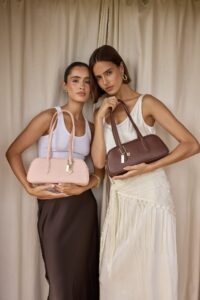 Two women pose side by side, each holding a structured handbag: blush pink on the left and brown on the right, in front of neutral curtains.